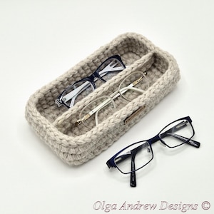 Puede incluir: Un soporte para gafas de ganchillo beige con dos compartimentos, que contiene tres pares de gafas. Las gafas son negras, blancas y con montura dorada. El soporte está sobre una superficie blanca. El texto "Olga Andrew Designs" está en la parte inferior.