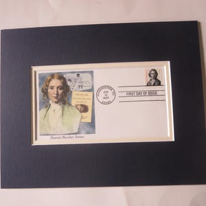 May include: A framed print featuring a postage stamp and a portrait of Harriet Beecher Stowe. The print includes the text "First Day of Issue" and a postmark from Washington, DC. The artwork is set against a white mount and a dark blue frame.