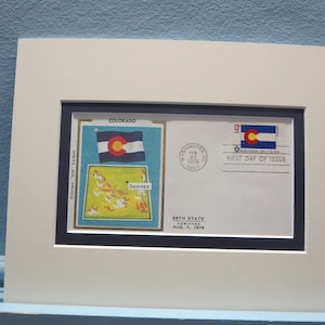 May include: A framed first day of issue envelope for the Colorado state commemorative stamp. The envelope features a blue and white state flag with a gold circle in the centre. The stamp depicts the state flag and the text "First Day of Issue". The envelope also includes a postmark from Washington, D.C. dated February 23, 1976.