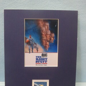 Puede incluir: Un póster enmarcado de la película "The Right Stuff" con un fondo azul y un grupo de astronautas en el centro. El póster tiene el título "The Right Stuff" y el lema "How the future began." Debajo del póster hay un pequeño sello con las palabras "Project Mercury".