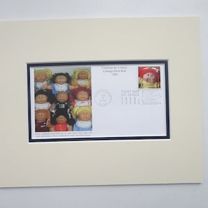 May include: A framed first day of issue envelope for the 1983 Cabbage Patch Kids stamp. The envelope features a photo of a group of Cabbage Patch Kids dolls and a stamp with a single doll. The text on the envelope reads "Celebrate the Century Cabbage Patch Kids 1983" and "First Day of Issue 1983".
