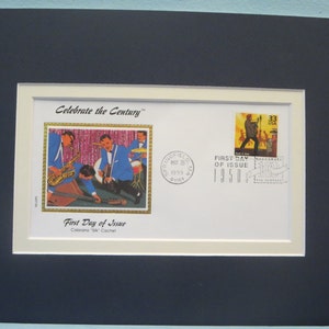 May include: A framed first day of issue envelope featuring a stamp with a band playing music. The envelope is white with a black border and a gold seal. The stamp is from 1950 and the envelope is from 1999. The text on the envelope reads "Celebrate the Century" and "First Day of Issue".
