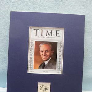 May include: A framed TIME magazine cover featuring Henry Ford, with a portrait and the magazine's logo. Below the magazine is a vintage 12-cent United States postage stamp. The frame is a dark blue colour.
