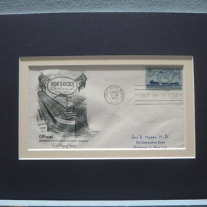 May include: Framed vintage envelope commemorating the Soo Locks Centennial, 1855-1955. The envelope features a black and white illustration of a ship and the text "First Day of Issue". Includes a blue postage stamp.