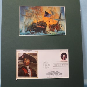 Puede incluir: Una impresión enmarcada de una pintura que representa una batalla naval. La pintura muestra dos barcos disparándose cañones el uno al otro. El barco de la izquierda está izando la bandera estadounidense. La impresión incluye un sello conmemorativo de John Paul Jones, el "Padre de la Armada Americana". El sello presenta un retrato de John Paul Jones y el texto "John Paul Jones, 1747-1792". El sello también incluye el texto "Todavía no he comenzado a luchar" y "Primer día de emisión".