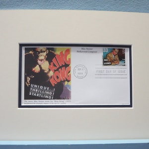 May include: A framed first day of issue stamp featuring a black and white image of King Kong from the 1933 film. The stamp is surrounded by a white border and a black border. The text "Max Steiner Hollywood Composer" is printed above the stamp. The text "First Day of Issue" is printed below the stamp. The text "UNIQUE..! THRILLING! STARTLING!" is printed below the image of King Kong. The text "The music Max Steiner wrote for "King Kong" (1933) highlighted the dramatic impact of the movie's action." is printed below the image of King Kong.