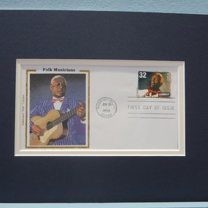 May include: Framed artwork featuring a vintage envelope and stamps. The envelope is stamped "Washington, DC, June 26, 1998" and has a stamp with a portrait of a man playing a guitar. The artwork is matted in white and framed in a dark blue frame.