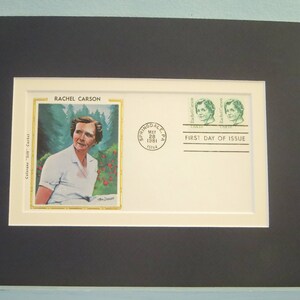 May include: A framed first day of issue envelope featuring a portrait of Rachel Carson, an American marine biologist, author, and conservationist. The envelope is white with a green stamp and the text "First Day of Issue".