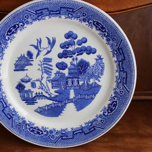 May include: A round, white ceramic plate with a blue Willow pattern design. The plate features a detailed landscape scene with buildings, trees, and birds. The rim has a decorative blue border. A classic piece for serving or display.