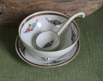 May include: A white porcelain soup tureen with a floral design and gold trim. The tureen has a matching saucer and a small ladle.
