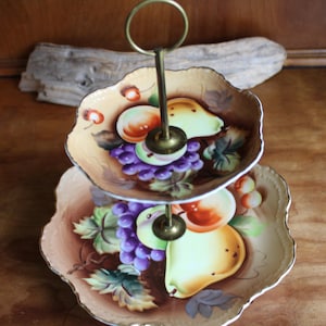 May include: Two-tiered serving tray with a gold handle. The plates are decorated with painted fruit, including grapes, pears, and peaches. The edges of the plates have a scalloped design and gold trim. A decorative piece for serving food.