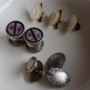 May include: A collection of vintage cufflinks and shirt studs in a white bowl. The cufflinks feature silver metal with purple and black enamel designs. Other studs are white and gold-toned, and silver with textured patterns.
