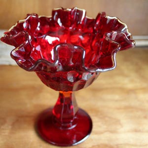 May include: A vintage ruby red glass pedestal bowl. The bowl has a ruffled rim with a gold trim. The bowl sits on a pedestal base. The glass has a glossy finish. The bowl is likely used for serving or display.