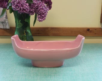 May include: A light pink ceramic serving dish with a boat-like shape. The dish has a wide, shallow bowl with raised handles on each end. A green glass vase with purple flowers is in the background.