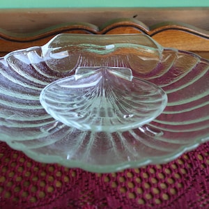 May include: A clear glass serving dish shaped like a seashell with three sections. The dish is on a red crocheted doily.