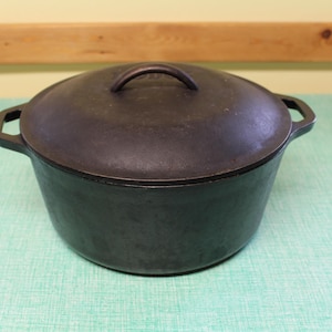 May include: A dark gray cast iron Dutch oven with a matching lid. The pot has two side handles and a loop handle on the lid. The pot is sitting on a light green surface.