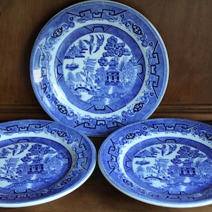 May include: Three blue and white ceramic plates with a classic Willow pattern. The plates feature a central landscape scene with pagodas, trees, and birds, surrounded by a decorative border. The plates are arranged on a wooden surface.