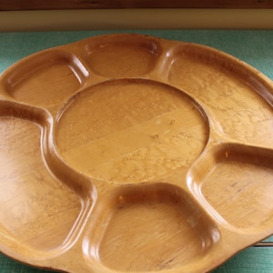 May include: A round, light brown wooden serving tray with multiple compartments. The tray has a central circular section surrounded by six curved sections, ideal for snacks or appetizers.