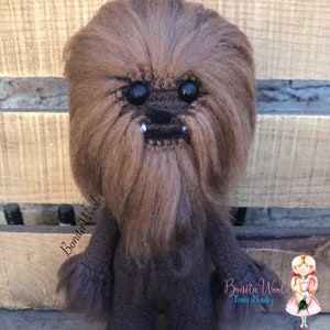 K&ouml;nnte beinhalten: Ein braunes und schwarzes, flauschiges Chewbacca-Pl&uuml;schtier mit gro&szlig;en schwarzen Augen und einem wei&szlig;en Bart. Das Spielzeug steht auf einer Holzoberfl&auml;che. Der Text "Bonita Wool!" ist auf der linken Seite des Bildes sichtbar.