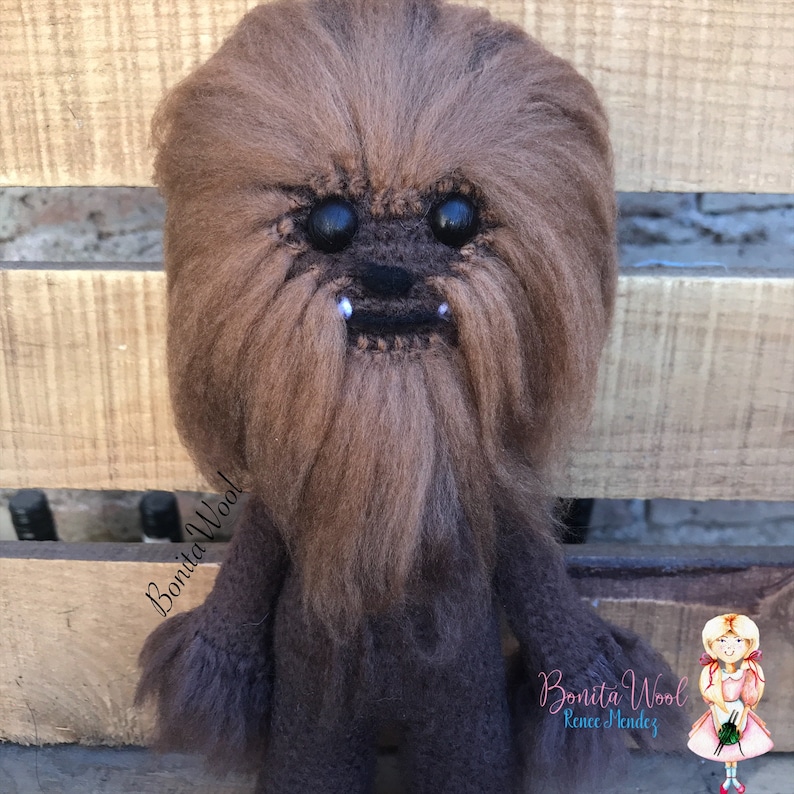 K&ouml;nnte beinhalten: Ein braunes und schwarzes, flauschiges Chewbacca-Pl&uuml;schtier mit gro&szlig;en schwarzen Augen und einem wei&szlig;en Bart. Das Spielzeug steht auf einer Holzoberfl&auml;che. Der Text "Bonita Wool!" ist auf der linken Seite des Bildes sichtbar.