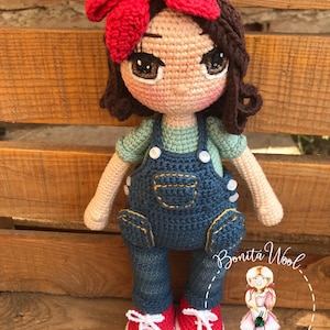 May include: A handmade crochet doll with brown hair, wearing a red bow, green shirt, denim overalls, and red shoes. The doll has large, expressive eyes and is posed against a wooden background. The doll is a unique, handcrafted item.