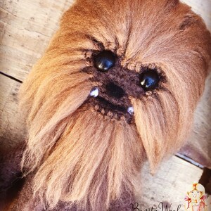 K&ouml;nnte beinhalten: Nahaufnahme eines braunen geh&auml;kelten Chewbacca-Charakters mit schwarzen Augen und wei&szlig;en Z&auml;hnen. Der Charakter ist aus Garn gefertigt und hat eine pelzige Textur. Der Hintergrund ist eine Holzoberfl&auml;che. Bonita Wool Renee Mendez steht in der rechten unteren Ecke des Bildes.