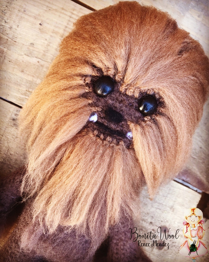 K&ouml;nnte beinhalten: Nahaufnahme eines braunen geh&auml;kelten Chewbacca-Charakters mit schwarzen Augen und wei&szlig;en Z&auml;hnen. Der Charakter ist aus Garn gefertigt und hat eine pelzige Textur. Der Hintergrund ist eine Holzoberfl&auml;che. Bonita Wool Renee Mendez steht in der rechten unteren Ecke des Bildes.