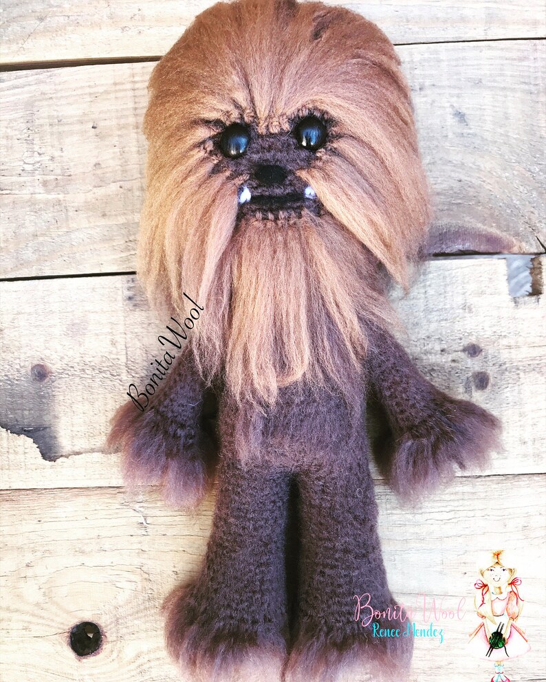 K&ouml;nnte beinhalten: Ein braunes, geh&auml;keltes Chewbacca-Pl&uuml;schtier mit schwarzen Augen und einem wei&szlig;en Bart. Das Spielzeug steht auf einer Holzoberfl&auml;che. Der Text "Bonita Wool!" ist auf dem Spielzeug geschrieben.