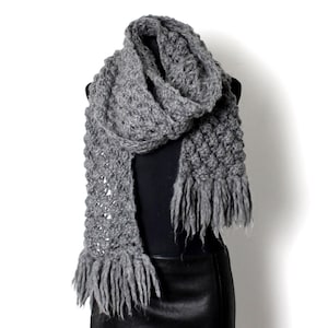 May include: A grey knitted scarf with fringe on a black mannequin. The scarf is made from chunky yarn and has a textured pattern.