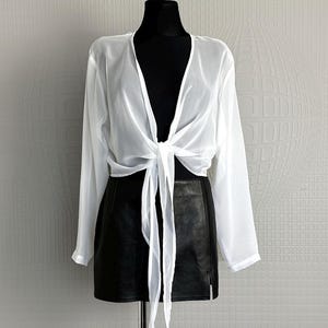 May include: A white long-sleeved blouse with a deep V-neckline and a front tie closure, paired with a black mini skirt. The blouse is made of a sheer, lightweight fabric, while the skirt features a smooth, solid black finish and a side zipper.