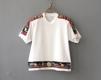 May include: White short-sleeved t-shirt with colorful patterned trim around the neckline and sleeves. The trim features a geometric design with blue, red, and yellow accents.