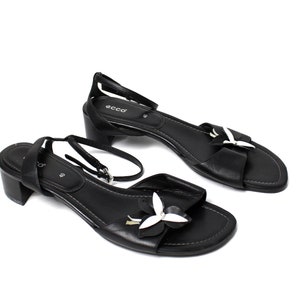 May include: A pair of black leather heeled sandals with a white flower accent. The sandals have a buckle closure and an ankle strap.