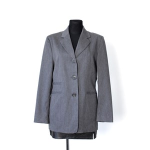 Pode incluir: Um blazer cinzento com lapela de entalhe, mangas compridas e fecho frontal de três botões. A peça inclui dois bolsos horizontais com debrum na parte da frente e é exibida num torso de manequim preto sobre um fundo branco.