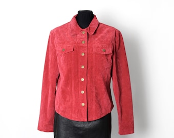 Puede incluir: Chaqueta de ante roja con cuello, dos bolsillos en el pecho y botones dorados. La chaqueta se exhibe en un maniquí negro, combinada con una falda negra. La chaqueta tiene mangas largas.