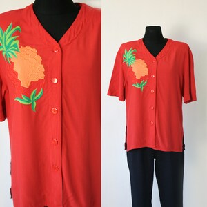 Può includere: Camicia rossa a maniche corte con bottoni e un ananas ricamato. L'ananas è verde, arancione e giallo.