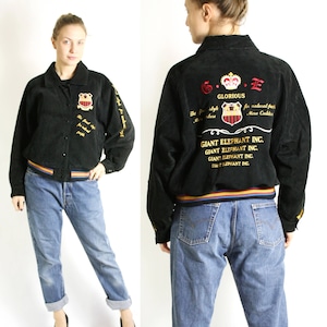 Peut inclure: Blouson noir en daim avec broderies dorées. La broderie comprend le texte "G.E. Glorious" et "Giant Elephant Inc." répété quatre fois. Le blouson a un col, des poignets et une ceinture côtelés.