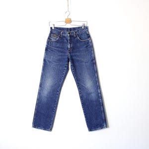 Pu&ograve; includere: Jeans blu in denim appesi a una gruccia di legno. I jeans hanno un classico design a cinque tasche, una chiusura con bottone e cerniera e una gamba dritta. Il denim ha un aspetto sbiadito e usurato.