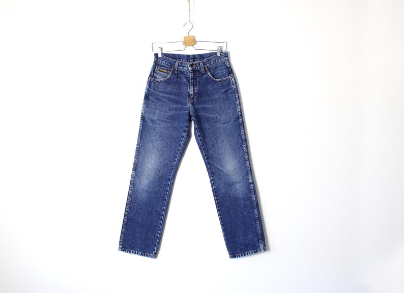 Pu&ograve; includere: Jeans blu in denim appesi a una gruccia di legno. I jeans hanno un classico design a cinque tasche, una chiusura con bottone e cerniera e una gamba dritta. Il denim ha un aspetto sbiadito e usurato.