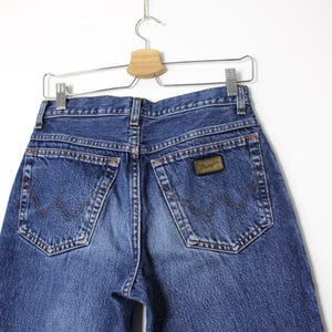 Pu&ograve; includere: Jeans Wrangler in denim blu, vista posteriore, appesi a una gruccia di metallo. I jeans hanno due tasche posteriori e una toppa in pelle marrone con il logo Wrangler. Il denim &egrave; sbiadito in alcune zone.