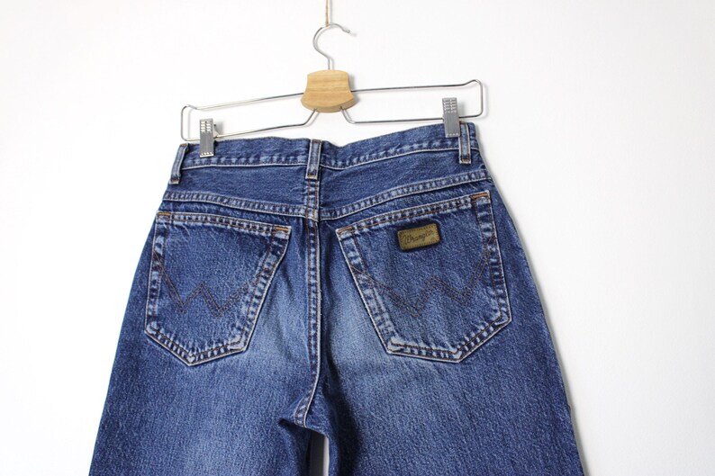 Pu&ograve; includere: Jeans Wrangler in denim blu, vista posteriore, appesi a una gruccia di metallo. I jeans hanno due tasche posteriori e una toppa in pelle marrone con il logo Wrangler. Il denim &egrave; sbiadito in alcune zone.