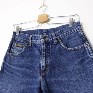 Pu&ograve; includere: Primo piano di jeans di denim blu appesi a un'appendiabiti. I jeans hanno un classico design a cinque tasche, una chiusura con bottone e cerniera e una piccola toppa con logo sulla tasca anteriore. Il denim ha un aspetto leggermente sbiadito.