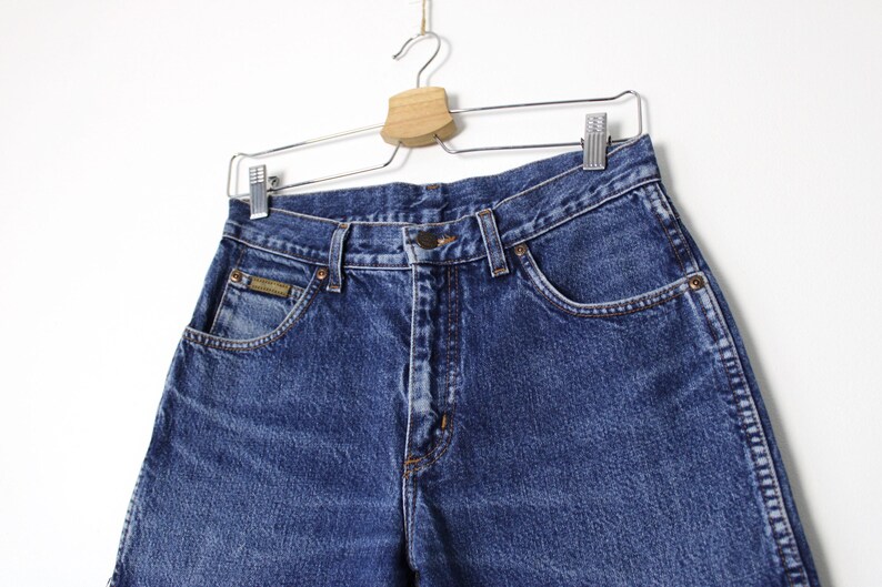 Pu&ograve; includere: Primo piano di jeans di denim blu appesi a un'appendiabiti. I jeans hanno un classico design a cinque tasche, una chiusura con bottone e cerniera e una piccola toppa con logo sulla tasca anteriore. Il denim ha un aspetto leggermente sbiadito.