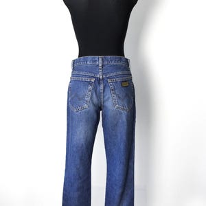 Pu&ograve; includere: Jeans in denim blu con un classico design a cinque tasche, esposti su un manichino nero. I jeans hanno un lavaggio sbiadito e una piccola toppa marrone sulla tasca posteriore. I jeans sono a gamba dritta.