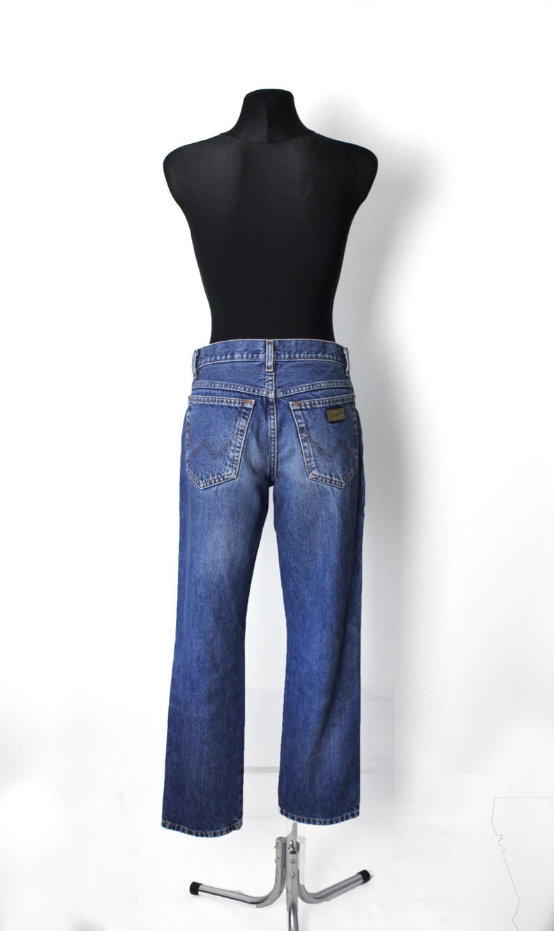 Pu&ograve; includere: Jeans in denim blu con un classico design a cinque tasche, esposti su un manichino nero. I jeans hanno un lavaggio sbiadito e una piccola toppa marrone sulla tasca posteriore. I jeans sono a gamba dritta.