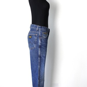 Pu&ograve; includere: Jeans in denim blu con una classica vestibilit&agrave; a gamba dritta. I jeans presentano un design tradizionale a cinque tasche, una chiusura con bottone e cerniera e una piccola toppa con logo sulla tasca posteriore. I jeans sono esposti su un manichino.