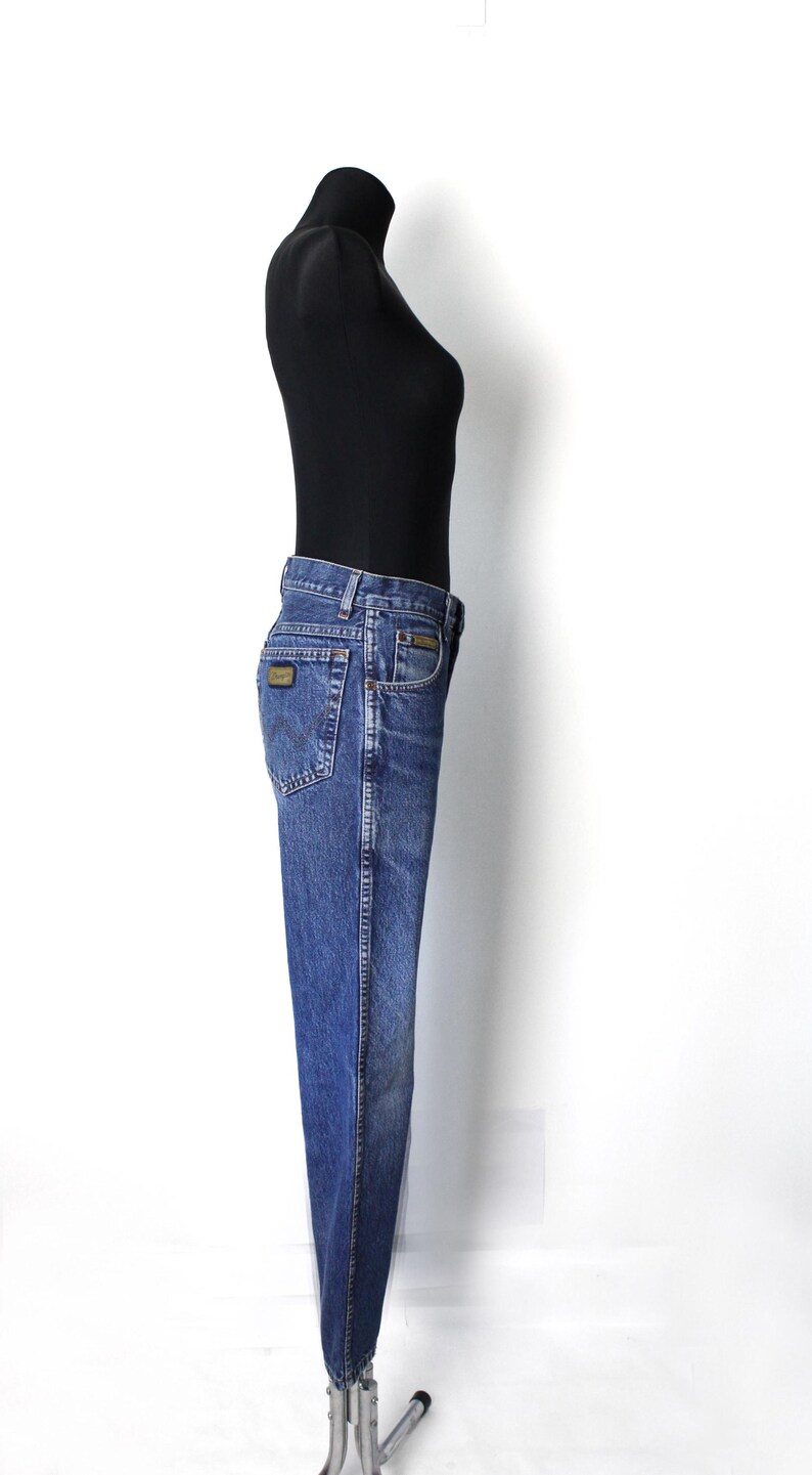 Pu&ograve; includere: Jeans in denim blu con una classica vestibilit&agrave; a gamba dritta. I jeans presentano un design tradizionale a cinque tasche, una chiusura con bottone e cerniera e una piccola toppa con logo sulla tasca posteriore. I jeans sono esposti su un manichino.