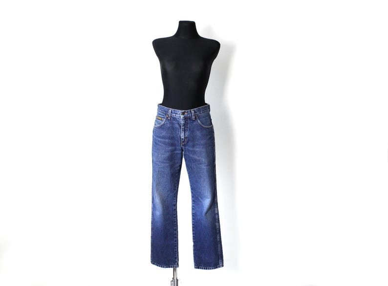 Pu&ograve; includere: Jeans di denim blu su un manichino nero. I jeans hanno una classica vestibilit&agrave; a gamba dritta, una chiusura con bottoni e cerniera e tasche. Il denim ha un lavaggio sbiadito, che gli conferisce un aspetto vintage. Il manichino &egrave; su sfondo bianco.