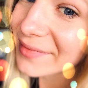 Op de afbeelding: Close-up van een gezicht met een kleine zilveren neusring. De persoon glimlacht en heeft blond haar. De achtergrond is wazig en heeft warme, wazige lichten.