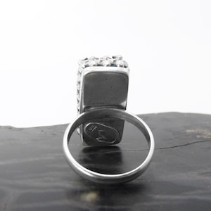 May include: A silver ring with a square-shaped stone set in a bezel setting. The ring is decorated with small beads around the stone. The ring is marked with a small symbol on the inside of the band.