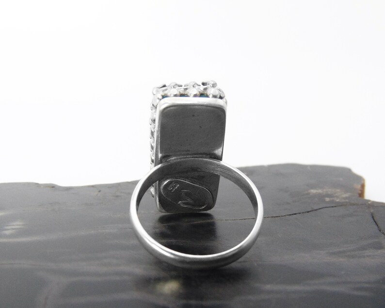 May include: A silver ring with a square-shaped stone set in a bezel setting. The ring is decorated with small beads around the stone. The ring is marked with a small symbol on the inside of the band.